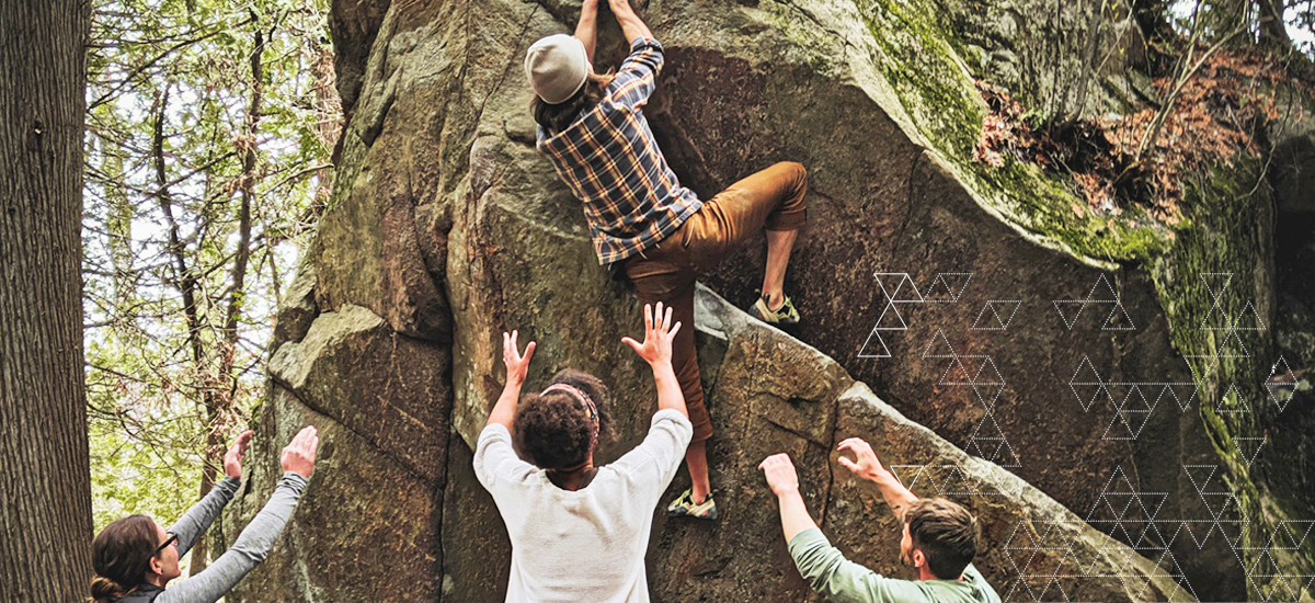 Man climbing rock with friends on the ground below with hands in the air to offer support